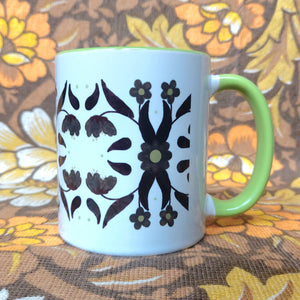 A white mug with a green handle and inside sits in front of a brown and white floral background. The mug features a symmetrical 70s inspired floral pattern in brown and green.