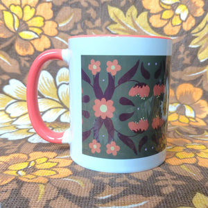 An orange and white mug sits in front of a brown and white floral background. The mug features a green rectangle with an orange and brown symmetrical leaf and flower pattern.