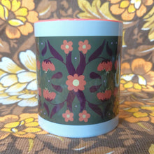 Load image into Gallery viewer, An orange and white mug sits in front of a brown and white floral background. The mug features a green rectangle with an orange and brown symmetrical leaf and flower pattern.
