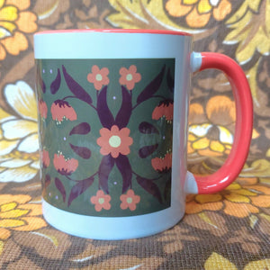 An orange and white mug sits in front of a brown and white floral background. The mug features a green rectangle with an orange and brown symmetrical leaf and flower pattern.