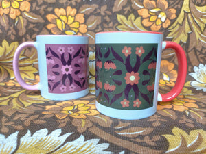 Two mugs with symmetrical floral patterns sits in front of a brown and white floral background. On the left is a pink mug and on the right and slightly closer to the camera is a green mug.
