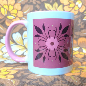 A white mug with pink handle sits on a brown white and yellow floral background. The mug features a pink and brown floral symmetrical pattern on a deep pink background. You can see it’s pink handle on the left.