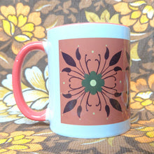 Load image into Gallery viewer, A white and orange mug sits on a brown and white floral background. The mug has an orange handle as well as a green and brown floral pattern on an orange background.
