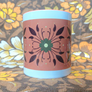 A white mug with a green and brown symmetrical floral pattern on an orange background sits in front of a white and brown floral background.