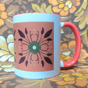 A white mug with an orange handle sits in front of a white and brown floral background. The mug features a rectangle of orange with a symmetrical green and brown floral pattern.