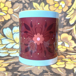 A white mug with a yellow handle and inside sits in front of a brown and yellow floral background. The mug features a brown rectangle across it with a dark orange and black floral pattern.