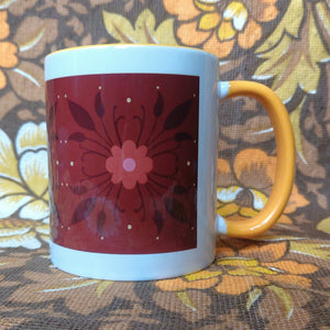 A white mug with a yellow handle and inside sits in front of a brown and yellow floral background. The mug features a brown rectangle across it with a dark orange and black floral pattern.