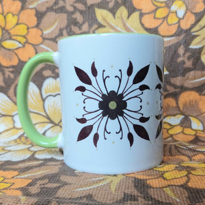 A white and green mug sits in front of a brown and white floral background. The mug has a green handle as well as a green and brown flower and leaf symmetrical parttern.