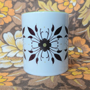 A white and green mug with a symmetrical floral pattern sits in front of a white and brown floral background.