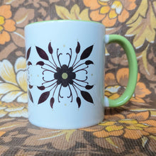 Load image into Gallery viewer, A white and green mug with a symmetrical floral pattern sits in front of a white and brown floral background. 
