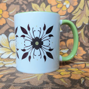 A white and green mug with a symmetrical floral pattern sits in front of a white and brown floral background. 