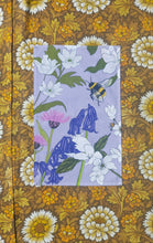 Load image into Gallery viewer, A lilac tea towel sits on a white, yellow and brown floral background. The tea towel features a range of different bee friendly flowers growing in from around he a[edges as well as a fluffy bumblebee!

