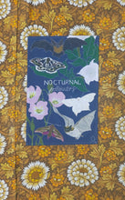 Load image into Gallery viewer, A deep blue tea glows featuring the words ‘Nocturnal Pollinators’ in white writing across the middle as well as a cou-or of bats, tombs and night flowering plants. Behind the tea towel is a warm brown, yellow and white floral patterned fabric.
