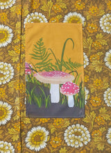 Load image into Gallery viewer, A warm yellow tea towel lies on warm brown, white and yellow floral fabric. The tea towel features a fly agaric, grass and fern design.
