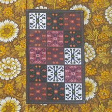 Load image into Gallery viewer, A dark brown tea towel sits on floral fabric which is yellow, white and brown. The tea towel features patterned floral tiles in a three by five formation, with four different colour ways across the design.
