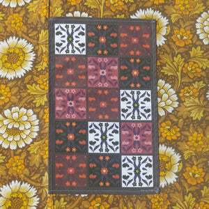 A dark brown tea towel sits on floral fabric which is yellow, white and brown. The tea towel features patterned floral tiles in a three by five formation, with four different colour ways across the design.