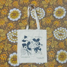 Load image into Gallery viewer, A natural off white tote sits in front of a white, brown and yellow floral patterned fabric. The bag features a black screen printed design of two different types of fungi and a flowering wild strawberry plant. There are also the names of the plants included around the outside of the design.
