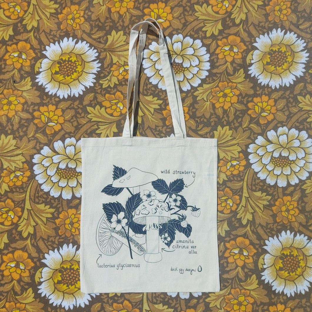 A natural off white tote sits in front of a white, brown and yellow floral patterned fabric. The bag features a black screen printed design of two different types of fungi and a flowering wild strawberry plant. There are also the names of the plants included around the outside of the design.