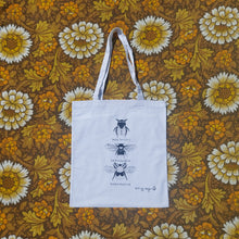 Load image into Gallery viewer, A white tote bag sits on a floral yellow, white and brown floral background. The tote bag features black and white illustrations of three bees in a line down the middle with their names underneath them.

