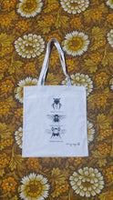 Load image into Gallery viewer,  A white tote bag sits on a floral yellow, white and brown floral background. The tote bag features black and white illustrations of three bees in a line down the middle with their names underneath them.
