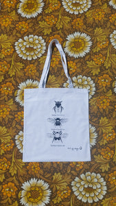  A white tote bag sits on a floral yellow, white and brown floral background. The tote bag features black and white illustrations of three bees in a line down the middle with their names underneath them.