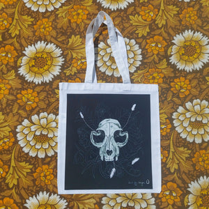 A white bag featuring a black square with a cat slkull in the middle with leafy fronds and fungi growing out from behind it. Behind the tea towel you can see a warm brown, yellow and white flora patterned fabric.
