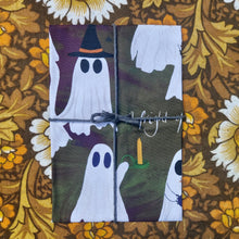 Load image into Gallery viewer, A deep purple and green tea towel featuring ghosts is folded and tied with grey macrame twine. Behind the tea towel you can see a brow, white and yellow floral patterned background.
