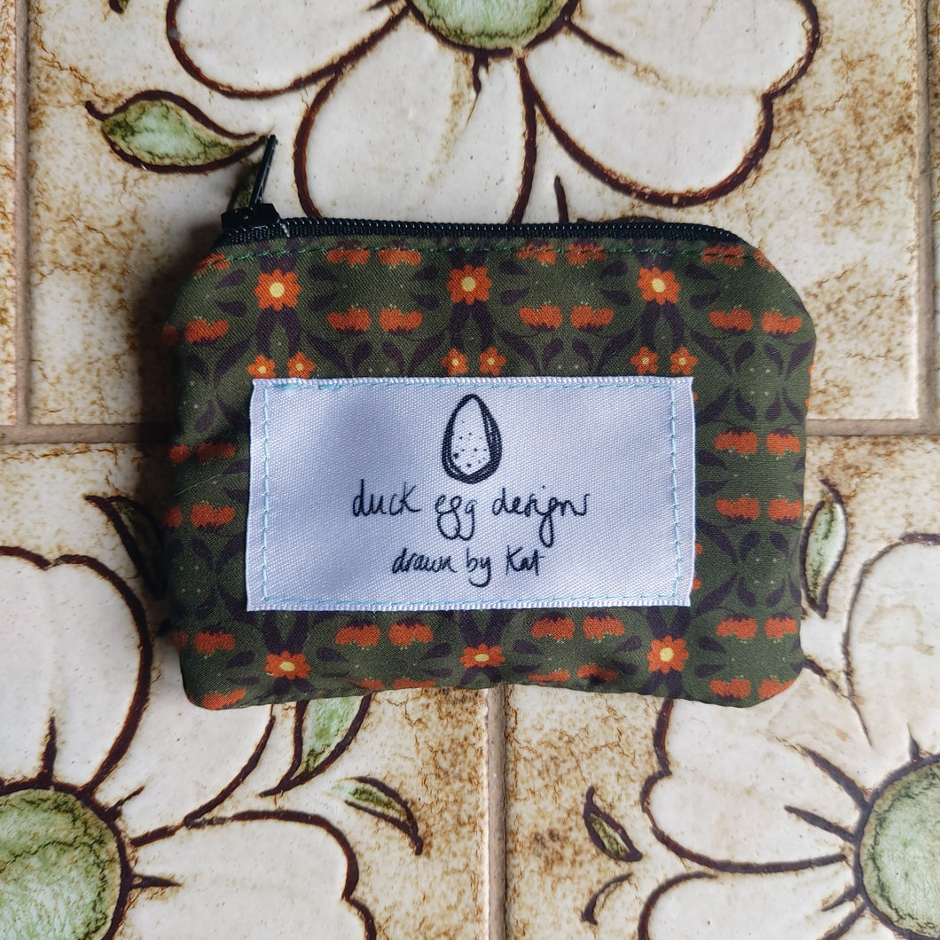 A green pouch with a black zip sits on a pale brown floral tile background. The pouch fabric has a symmetrical repeating floral pattern across it in orange and dark brown, with a white label featuring the Duck Egg Designs logo.