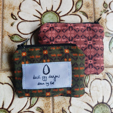 Load image into Gallery viewer, A green pouch with an orange and brown floral pattern sits on top of a pink pouch with the same pattern in light pink and brown. You can see a white label on the green pouch with the Duck Egg Designs logo. Behind the pouches you can see light brown tiles with white and green flowers on them.
