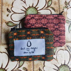 A green pouch with an orange and brown floral pattern sits on top of a pink pouch with the same pattern in light pink and brown. You can see a white label on the green pouch with the Duck Egg Designs logo. Behind the pouches you can see light brown tiles with white and green flowers on them.