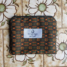 Load image into Gallery viewer, A dark green pouch with a symmetrical floral print in orange and dark brown sits on a pale brown floral tile background. The pouch features a white label with the Duck Egg Designs logo in black.
