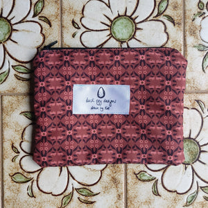 A dusty pink pouch with  a repeating pale pink and brown symmetrical repeating pattern. The pouch features a white label with the Duck Egg Designs logo on it and has a black zip. Behind the pouch are pale brown tiles with white and green flowers on them.