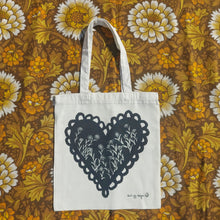Load image into Gallery viewer, A white bag sits on a retro floral patterned brown, yellow and white fabric. The tote bag features a black heart design with white daisy style flowers growing across it from the bottom edges; to the bottom right of the design you can see the Duck Egg Designs logo.
