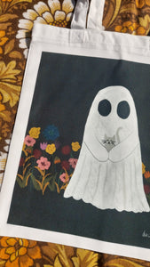 A close up of the bag, showing the detail of the colourful flowers as well as a closer look at the ghost holding the happy grey kitten. The design sits on a dark grey square on the white tote bag, and to the left of the bag you can see a retro floral pattern in white, yellow and brown.