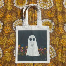 Load image into Gallery viewer, A white bag sits on a retro floral brown, orange and white fabric. The bag features a dark grey square with a ghost holding a grey kitten in the middle of it. To the either side of the ghost are flowers in a range of different colours, growing from the bottom of the square. In the bottom right is a white Duck Egg Designs logo.
