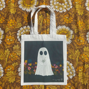 A white bag sits on a retro floral brown, orange and white fabric. The bag features a dark grey square with a ghost holding a grey kitten in the middle of it. To the either side of the ghost are flowers in a range of different colours, growing from the bottom of the square. In the bottom right is a white Duck Egg Designs logo.