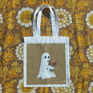 A white tote bag sits on a retro floral brown, white and yellow patterned background. The tote features a golden yellow rectangle with a ghost hugging a bunch of flowers. To the bottom right of the square is the Duck Egg Designs logo in white.