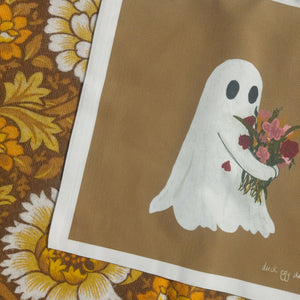 A close up of the bottom right of the bag, showing the flower holding ghost in more detail. To the left of the bag you can see a retro floral patterned background in brown, white and yellow.