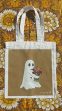Load image into Gallery viewer, A white tote bag sits on a retro floral brown, white and yellow patterned background. The tote features a golden yellow rectangle with a ghost hugging a bunch of flowers. To the bottom right of the square is the Duck Egg Designs logo in white.
