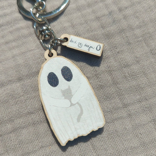 A white ghost keyring holding a grey kitten sits on a light grey fabric background. 