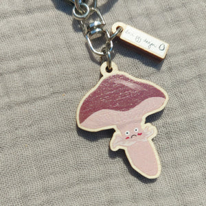 A close up of the keyring focusing on the fungi charm. The fungi has a light brown, almost pink stem with a little unhappy face. The cap of the fungi is a warm deep brown and you can also see the white and black Duck Egg Designs logo charm above it.