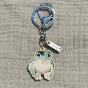 A green and pink frog keyring with a split ring and a lobster clasp sits on a light grey fabric background. Just above the frog you can see a second mini charm with the white and black Duck Egg Designs logo.