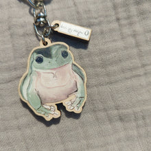 Load image into Gallery viewer, A green and pale pink happy frog keyring sits on a light grey fabric. Above the frog charm you can see a small rectangle charm with the white and black Duck Egg Designs logo.
