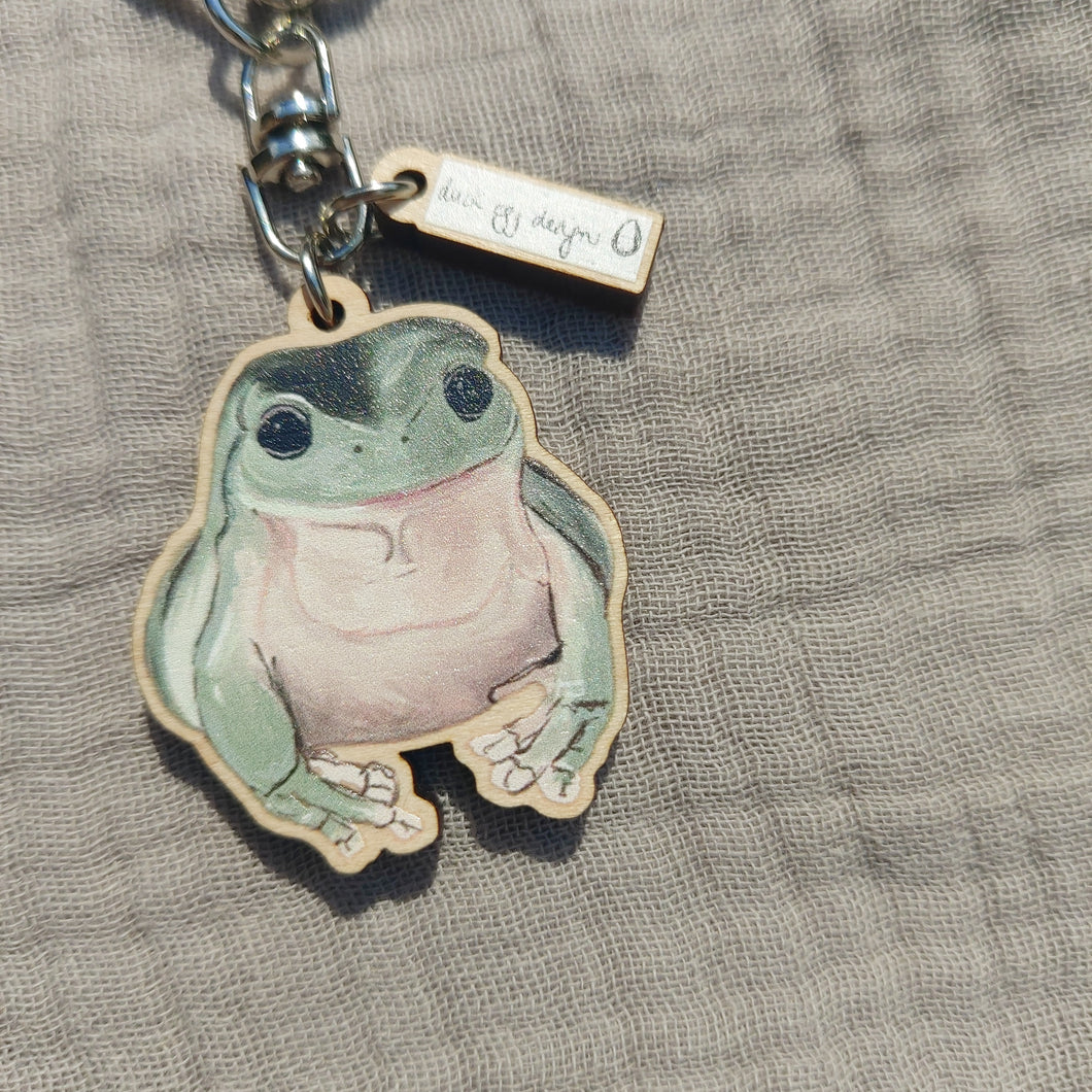 A green and pale pink happy frog keyring sits on a light grey fabric. Above the frog charm you can see a small rectangle charm with the white and black Duck Egg Designs logo.