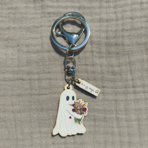A ghost keyring with a split ring and a lobster clasp sits on a light grey background. The ghost is white with black eyes and hugs a bunch of richly coloured flowers in red, dusky pink and mustard yellow. 