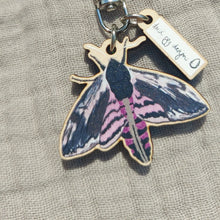 Load image into Gallery viewer, A pink and black moth keyring sits on a grey fabric background. The keyring has a white min charm with a Duck Egg Designs logo.
