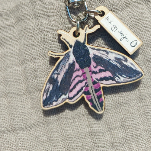 A pink and black moth keyring sits on a grey fabric background. The keyring has a white min charm with a Duck Egg Designs logo.