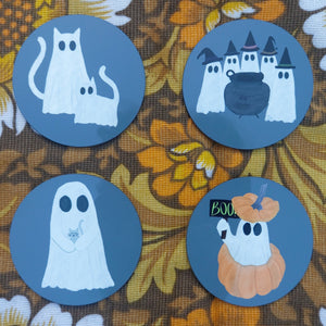 Four grey coasters sit on a warm brown floral retro patterned fabric. He coasters feature different ghost designs from L to R, top to bottom - two ghost cats, a coven of ghost witches around a cauldron, a ghost holding a grey kitten and a ghost popping out of a pumpkin with a little sign that reads BOO!.