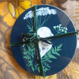 A set of black coasters sits on a floral patterned brown retro fabric. The coasters are tied together with a velvety green ribbon, and you can see the raven skull and hemlock design on the top coaster.