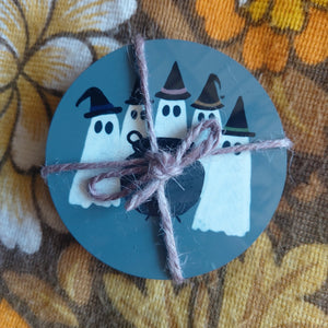 A full and packaged coaster set tied together with dusky purple string. You can see the ghost coven design on the top circular grey coaster - with a group of ghosts wearing witches hats standing around a cauldron. Behind the coaster set is a floral patterned background in brown and orange and white.